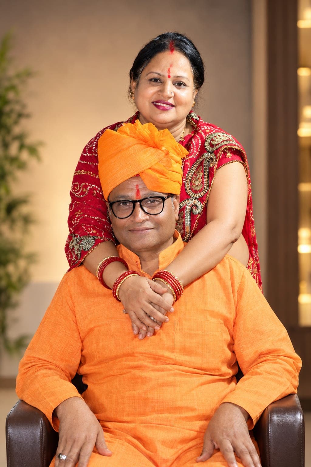 Shalu Mishra in a red saree, standing with her hands on the shoulders of a man in an orange kurta and turban seated in front of her